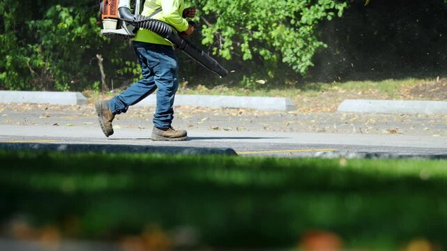 Worker using a leaf blower to clear fallen leaves and debris along edge of street or parking lot. Dust and dry leaves swirl into the air as the blower operates.