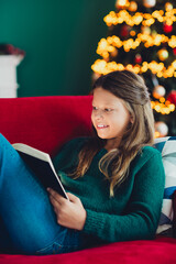 Naklejka premium A funny cozy scene of a girl reading a book beside a glowing Christmas tree during winter holidays at home