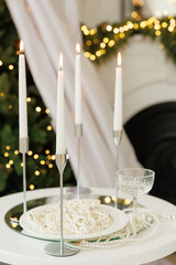 Elegant close-up of four lit white taper candles in silver holders, a plate of pearls, and a champagne glass on a white table with bokeh lights.