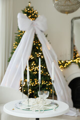 A white table with pearls, a champagne glass, and four candles, set against a blurred background of a Christmas tree draped in white sheer fabric.