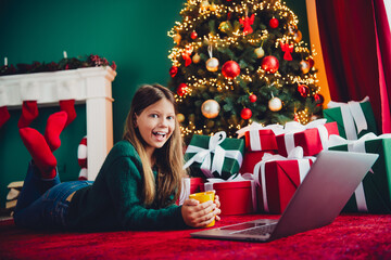 funny cozy christmas moment girl with laptop by christmas tree surrounded by gifts and twinkling lights at home