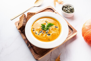 Hot spicy pumpkin soup with seeds, oregano leaves and bread in white ceramic plate on light marble background close up. Healthy vegan food concept.