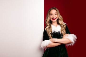 Festive glamorous woman in green velvet fur trimmed dress smiles arms crossed against a split white...