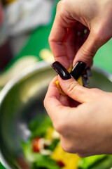 Fototapeta premium A close-up shot of a person's hands with dark nail polish, adding a piece of yellow food to a fresh green salad in a shiny metal bowl during food preparation.