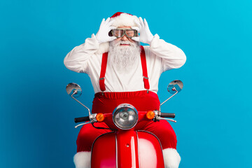Santa on a bright red scooter poses joyfully wearing hat suspenders and gloves in a blue studio for...