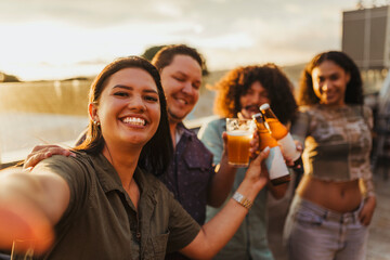 Happy friends taking a selfie while toasting with beer on a rooftop at sunset