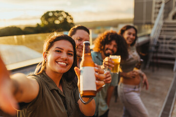 Smiling friends taking a selfie while toasting with beer bottles at a rooftop party