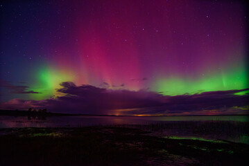 Colorful aurora borealis with vivid green and magenta lights illuminating the night sky above a calm lake and dark clouds.