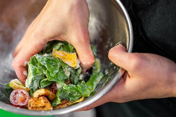 A close-up shot of hands mixing a fresh, healthy Caesar salad in a metal bowl. The salad contains romaine lettuce, croutons, chicken, and cherry tomatoes with creamy dressing