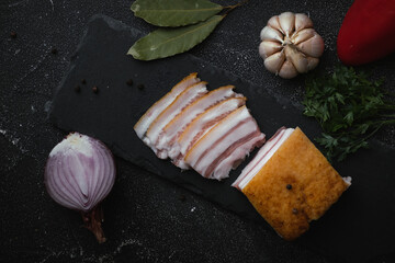 Salted lard bacon sliced on the black concrete background decorated with bay leaf,  garlic and onion. Pork fat meat as healthy food. Top view. Traditional Ukrainian food