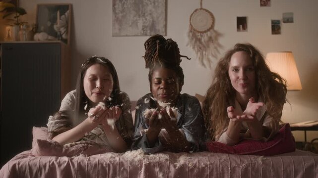 Medium portrait of three diverse girlfriends, Caucasian, Asian, African American, lying on bed, dressed in pajamas, blowing off handfuls of feathers after pillow fight, looking at camera, laughing