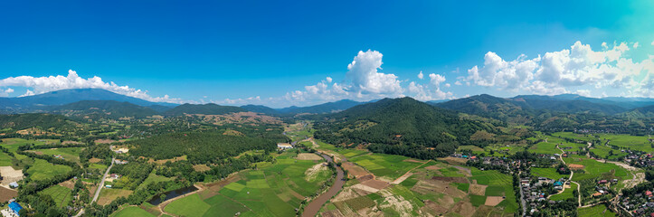 Aerial of lush rice fields with wooden boardwalk, flying high toward green mountains and blue sky white clouds near rural countryside. Peaceful, natural daytime landscape