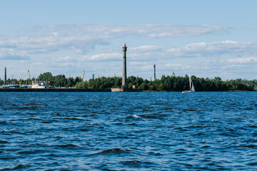 Obraz premium Panoramic view of a deep blue sea with a lighthouse and small sailing boat near the green wooded shore under a cloudy summer sky