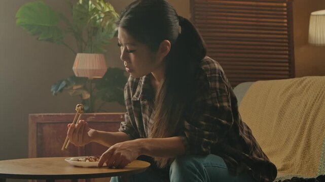 A Korean woman is sitting at a table enjoying traditional Asian cuisine with chopsticks in a cozy home setting.