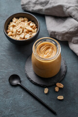 Creamy peanut butter in a jar on a black plate on a dark background with a scattering of roasted nuts