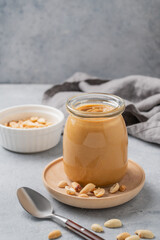Creamy peanut butter in a jar on a wooden plate on a blue background with a scattering of roasted nuts