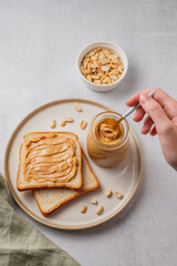 A hand holds a spoon with creamy peanut butter in a jar on a plate with toast and roasted nuts