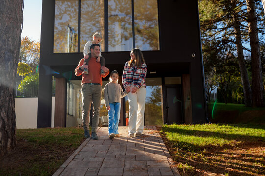 Happy family of four walking hand in hand along a paved path in front of their modern suburban home, enjoying a sunny autumn day and family togetherness