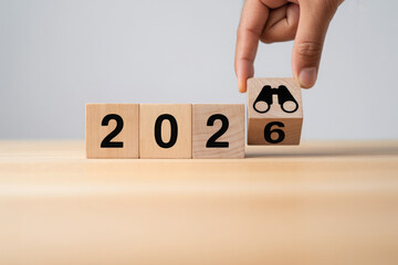 Hand changing wooden blocks from 2025 to 2026 symbolizing future vision and business planning with a binoculars icon for strategic foresight