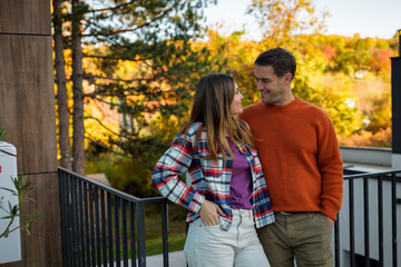 Fototapeta premium Happy couple standing close, looking into each other's eyes and smiling, sharing a romantic moment outdoors on a balcony with vibrant autumn trees in the background