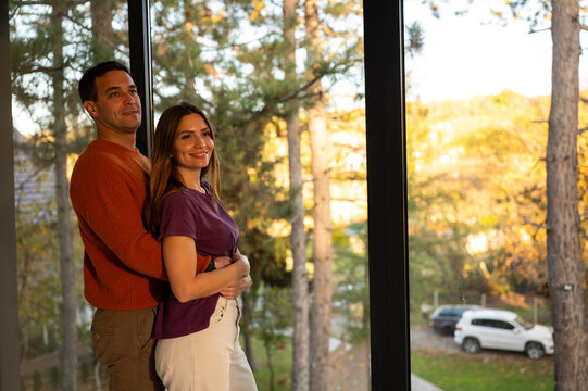 Happy couple embracing, looking out large window at colorful autumn trees, enjoying serene home environment and contemplating future lifestyle together