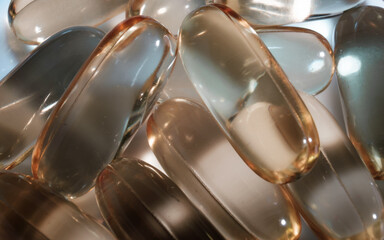 Naklejka premium Zbliżenie na stos owalnych, przezroczystych kapsułek z suplementami diety, o bursztynowej barwie. Kapsułki są ułożone losowo, a ich lśniąca powierzchnia odbija światło.