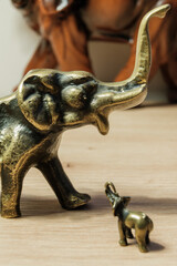 Dwa mosiężne słonie na drewnianym stole. Dwie figurki słoni, wykonane z mosiądzu, stoją na drewnianym blacie. Większy słoń patrzy na mniejszego, który unosi trąbę w górę. 