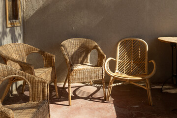 Three vintage wicker and rattan chairs sitting on a porch or patio against a plain wall, illuminated by strong afternoon sunlight and casting long, pronounced shadows.
