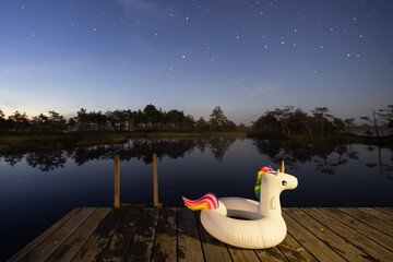 Inflatable unicorn float on a wooden dock under a clear, starry night sky reflected in a calm dark lake.