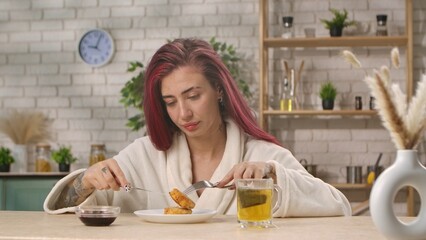A woman in a white robe sits at a kitchen table eating breakfast with herbal tea in a cozy home setting. Breakfast in the home kitchen, the morning ritual of eating.