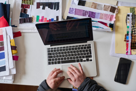 Fashion designer working on laptop surrounded by fabric swatches, creating new collection, top view, fashion industry concept, design process, creative workspace