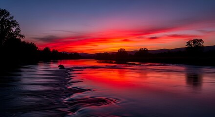 River reflects stunning fiery sunset, with trees silhouetted against vibrant red and orange sky