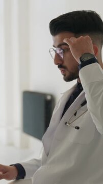 Vertical video. Tired and overwhelmed young physician rubbing his eyes at his desk in a bustling hospital, feeling the effects of burnout and fatigue from the rigors of the demanding medical field