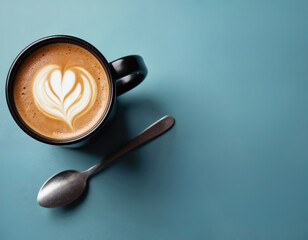 Top view close up of mug with heart latte art and spoon on blue background. Aroma coffee drink on cafe tabletop. Morning coffee break for barista. Sweet refreshment.