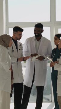 Vertical video. Multicultural medical team in hospital corridor reviewing records and discussing patient treatment plans, collaborating professionally