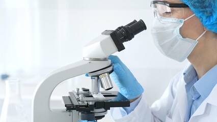 Female scientist analyzing research sample through microscope, wearing protective lab gear for medical investigation. Laboratory and science concept