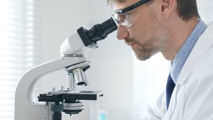 Adult man scientific researcher analyzing sample through microscope, wearing protective gear in high tech laboratory environment. Medicine and science concept