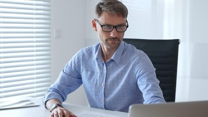 Male adult accountant wearing blue sleek shirt and black glasses is concentrating while working at the desk in modern office, demonstrating professionalism. Business people, taxes and audit concept