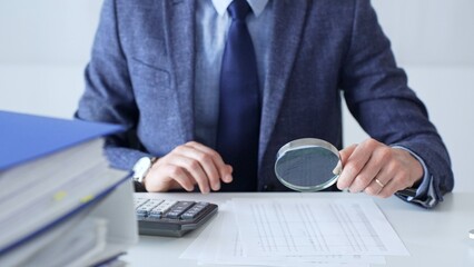 Professional accountant is examining financial documents, magnifying glass in hand, calculator...