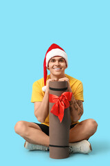 Obraz premium Young happy man in Santa hat sitting with aerobic mat on blue background