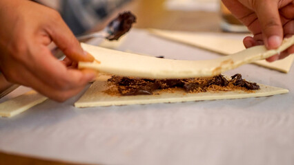 Close-up of hands folding croissant dough filled with chocolate spread and brown sugar at home kitchen, step of baking preparation