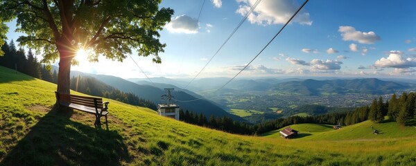 Green hill with lone tree and bench overlooks valley town. Chairlift station on grassy slope below blue sky with fluffy clouds. Sunny day with scenic mountain landscape.
