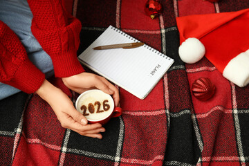 Woman holding cup of coffee, notebook with empty to do list and Santa hat on red checkered plaid