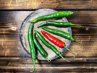 glossy green chili peppers with a single contrasting red chili in the center, all placed on a rustic wooden cutting board against a dark wooden surface.