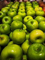 A dense, close-up view of fresh green apples (Granny Smith or similar) stacked in rows on a grocery shelf, featuring varying shades of bright green and yellow-green.