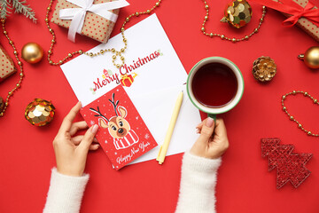 Female hands with greeting cards, cup of tea, Christmas decorations and gifts on red background