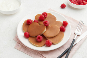 A heap of heart shaped pancakes on the white plate with raspberries on the white background