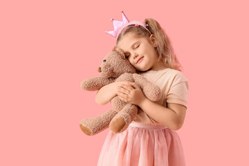 Fototapeta premium Adorable little girl with pink princess crown cuddling soft teddy bear toy on solid pink background, her eyes are closed in contentment, showing a moment of comfort and security