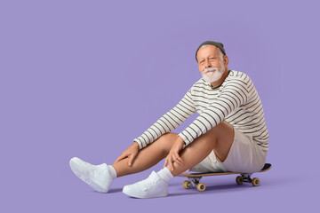Fototapeta premium Senior man sitting on skateboard against lilac background