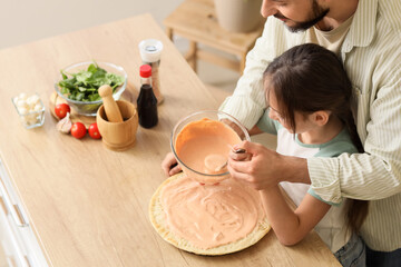 Father with his daughter applying sauce on pizza dough in kitchen, top view